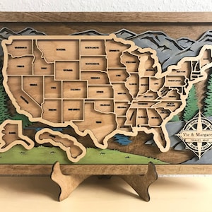 May include: A wooden map of the United States with each state outlined. The map is framed in a brown wood frame and has a compass rose in the lower right corner. The compass rose has the text "Vic & Margaret's Travel Map" on it.
