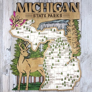 May include: A wooden map of Michigan State Parks with a deer silhouette in the lower left corner. The map is outlined in brown and features the names of each state park in white lettering.
