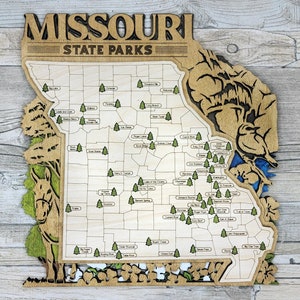 May include: A wooden map of Missouri State Parks with the names of each park printed on the map. The map is cut out in the shape of the state of Missouri and has a brown wood finish. The map is decorated with small green trees and a blue river.