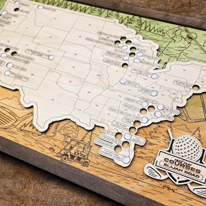 May include: Wooden golf course map of the United States, featuring a detailed laser-cut design. The map includes labeled golf courses and decorative golf-themed illustrations. The map is set in a wooden frame.