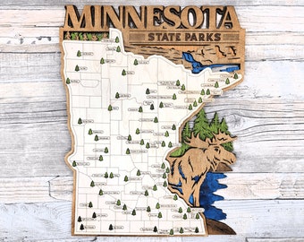 Può includere: Una mappa in legno del Minnesota con il testo "MINNESOTA STATE PARKS" in alto. La mappa è decorata con piccoli alberi verdi e il contorno di un alce. La mappa è su uno sfondo in legno chiaro.