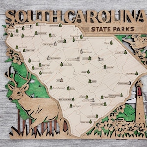 May include: Wooden map of South Carolina State Parks. The map is light brown with green and brown accents, featuring a deer, lighthouse, and floral details. The text "SOUTH CAROLINA STATE PARKS" is carved at the top, with park names labeled throughout.