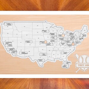 May include: A light-colored wooden map of the United States featuring state outlines and baseball icons representing various professional teams. A decorative emblem with crossed bats and a baseball sits in the corner with the text TEXT HERE.