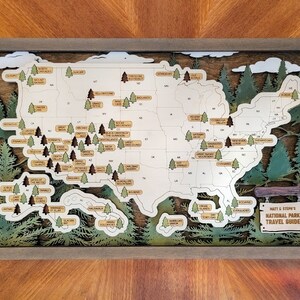 May include: A wooden map of the United States with a laser-cut design featuring national parks. The map is framed in a brown wood frame and includes a sign that reads "Matt & Steph's National Park Travel Guide."