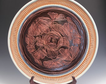 May include: A large ceramic plate with a brown glaze and a decorative design of stylized flowers in a copper color. The plate has a wide rim with a textured pattern in brown and white.