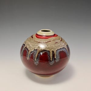 May include: A small, round ceramic vase with a red glaze and a white rim. The glaze has a mottled effect, with streaks of brown and white.