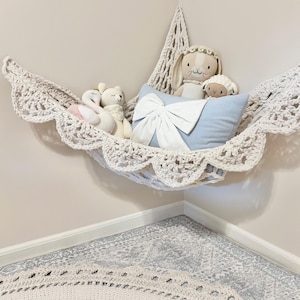 Puede incluir: Una hamaca de ganchillo color crema con un borde festoneado, colgada en una esquina. En el interior hay animales de peluche: un cisne, un oso, un conejito y un cordero, junto con una almohada azul claro con un lazo blanco. La hamaca está suspendida del techo.