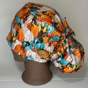 May include: A scrub cap featuring a vibrant pattern of orange, white, and teal pumpkins with green leaves and berries. The fabric head covering includes a round wooden button on the side and a gathered back section with ties for an adjustable fit.