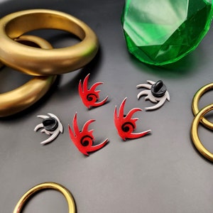 Könnte beinhalten: Fünf Emaille-Pins mit einem stilisierten roten und weißen Igel-Design. Die Pins befinden sich auf einer dunkelgrauen Oberfläche mit goldenen Ringen und einem grünen Edelstein.