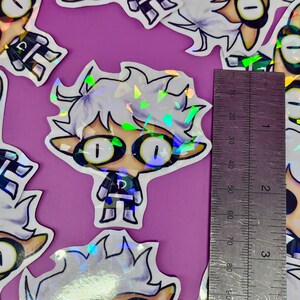 May include: Holographic stickers featuring a cartoon character with white hair and large eyes. The character is wearing a black and blue outfit. A ruler shows the stickers are approximately 3 inches in height. The stickers are on a purple background.