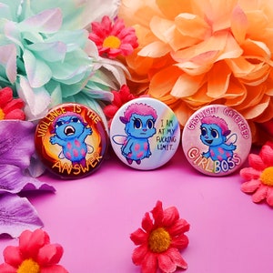Op de afbeelding: Drie ronde button badges met kleurrijke cartoonfiguren en tekst. De eerste badge zegt "Violence is the answer" met een blauw personage met rode ogen. De tweede badge zegt "I am at my fucking limit." met een blauw personage met roze haar. De derde badge zegt "Gaslight Gatekeep Girlboss" met een blauw personage met roze haar.