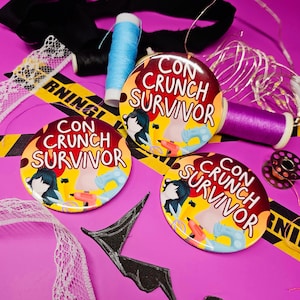 Könnte beinhalten: Drei runde Ansteckbuttons mit dem Text CON CRUNCH SURVIVOR in weißen Buchstaben. Das Design zeigt Illustrationen einer Nähmaschine, einer Heißklebepistole und einer Schneiderpuppe auf gelb-rotem Grund mit einer stilisierten Figur.