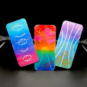 May include: Three rectangular art pieces with colorful abstract designs. One features eyes, another a sunset with stars, and the third has vertical lines and stars. The pieces are painted in shades of blue, pink, orange, and green.