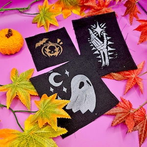 May include: Three black fabric patches with white and silver designs. The patches feature a ghost, a skeleton hand, and a pumpkin with a bat.