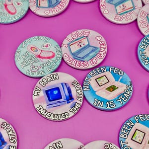 May include: A collection of round, colorful buttons with retro technology graphics and text. Designs include a flip phone, a laptop, a computer monitor, and a handheld gaming device. Text includes phrases like "I love dress up games" and "My brain inherited the static."