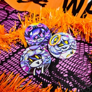 May include: Three circular pin-back buttons featuring stylized anime-inspired character illustrations. The designs showcase characters with purple and white hair, vibrant yellow eyes, and heart motifs, rendered in a glossy finish with bold outlines.