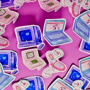 May include: A collection of colorful stickers featuring various 2000s tech items, including a pink Game Boy Advance, a blue laptop with the text "GirlDressUp.com", a blue DVD player, a pink flip phone, and a blue computer monitor. The stickers are on a pink background.