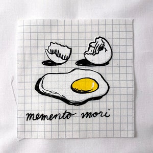May include: Black and white illustration of a broken egg, a cracked egg shell, and a fried egg on a gridded background. The text "memento mori" is written below the image.