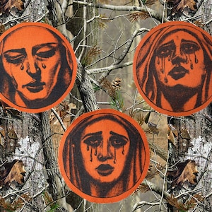 May include: Three round orange patches with black and white images of a woman's face with tears running down her cheeks. The woman is wearing a head covering.