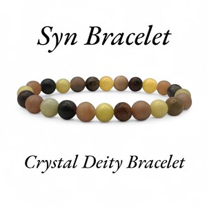 May include: A beaded bracelet with a variety of earth-toned beads, including brown, black, yellow, and beige. The bracelet is curved and displayed against a white background. Text on the image reads "Syn Bracelet" and "Crystal Deity Bracelet."