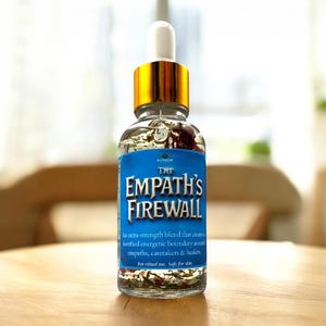 May include: A clear glass bottle with a gold cap and white dropper, labeled "THE EMPATH'S FIREWALL." The bottle contains a liquid with visible herbs and flowers. The label is blue with white text. The bottle is on a wooden surface.