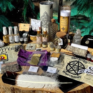 May include: A collection of witchy supplies on a wooden surface, including candles, herbs, crystals, and a feather. The text "Protected & Blessed by Witchcraft" is visible on a black cloth.