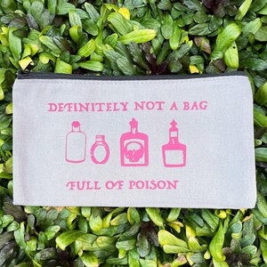 May include: A light gray zippered pouch with the text "DEFINITELY NOT A BAG" and "FULL OF POISON" in pink. The pouch features pink illustrations of potion bottles. The background is a bed of green leaves.