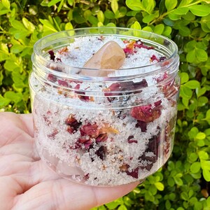 May include: A clear glass jar filled with white bath salts and dried rose petals. A small, light brown crystal is on top of the bath salts.