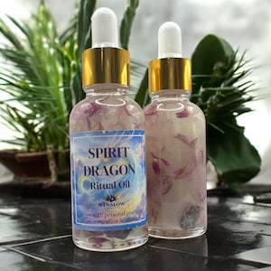 May include: Two glass bottles of Spirit Dragon Ritual Oil with gold-colored caps and white droppers. The bottles contain pink flower petals and a blue label with the text "SPIRIT DRAGON Ritual Oil". The background includes green plants.