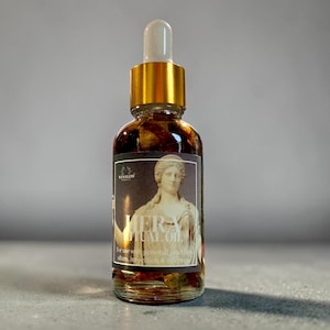 May include: A clear glass dropper bottle filled with amber liquid and botanical pieces. The label features a classical statue bust, the text WINSLOW, HERA RITUAL OIL, and For use with personal practices, altars, invocation & offerings.