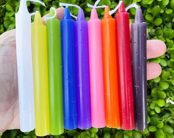 May include: A collection of eight colorful, slender candles in a rainbow of hues, including white, yellow, green, blue, purple, pink, orange, red, and dark purple. Each candle has a white wick. The candles are arranged in a row against a backdrop of green foliage.