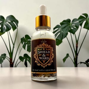 May include: A clear glass dropper bottle with a gold cap and a dark brown label featuring a gold shield emblem. The text reads Winslow Shield & Ward for ritual work or topical skin use. Dried botanical elements are visible at the base.