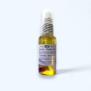 May include: A clear glass travel-size bottle of Crystodorant, with a white cap and a label that reads "Ancestral Awakening." The bottle contains a yellow liquid with visible particles. The net weight is 1 oz.