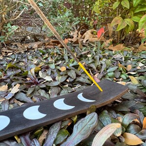 Puede incluir: Un porta incienso de madera negra con cinco formas de media luna blancas. Un palo de incienso amarillo est&aacute; ardiendo en el porta incienso.