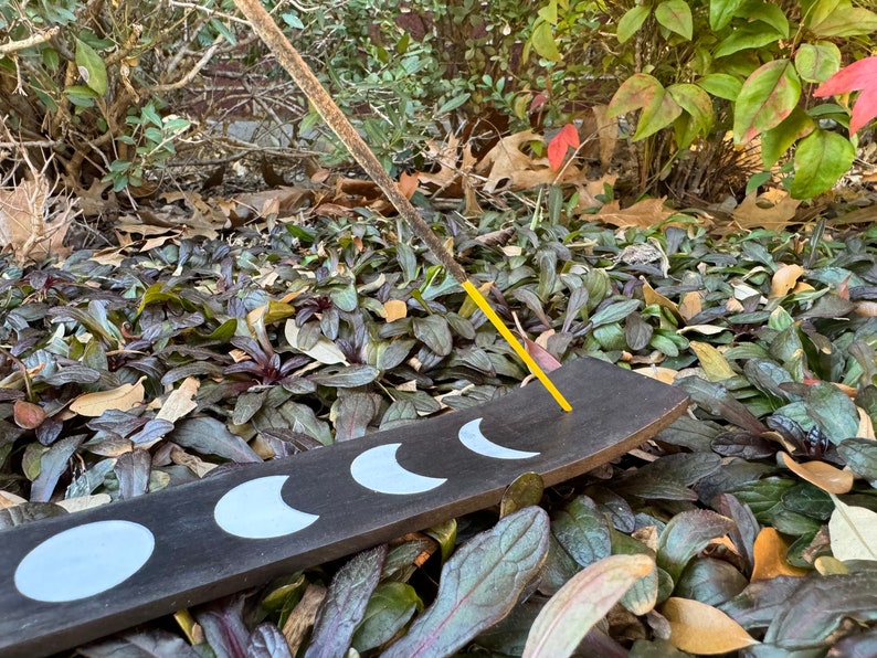 Puede incluir: Un porta incienso de madera negra con cinco formas de media luna blancas. Un palo de incienso amarillo est&aacute; ardiendo en el porta incienso.