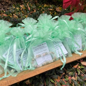 May include: A collection of small green organza bags filled with colorful gemstone bracelets. Each bag has a small card with the name of the bracelet and the type of gemstone used. The text on the cards reads "In Child Work Handstrung Crystal Bracelet - Dalmatian Jasper" and "Protection Anustp Stal B Ger".