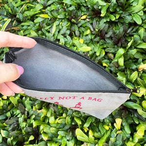 May include: A canvas zippered pouch with the text "DEFINITELY NOT A BAG" in red. The pouch is open, revealing a gray interior. The background is a dense bed of green foliage.