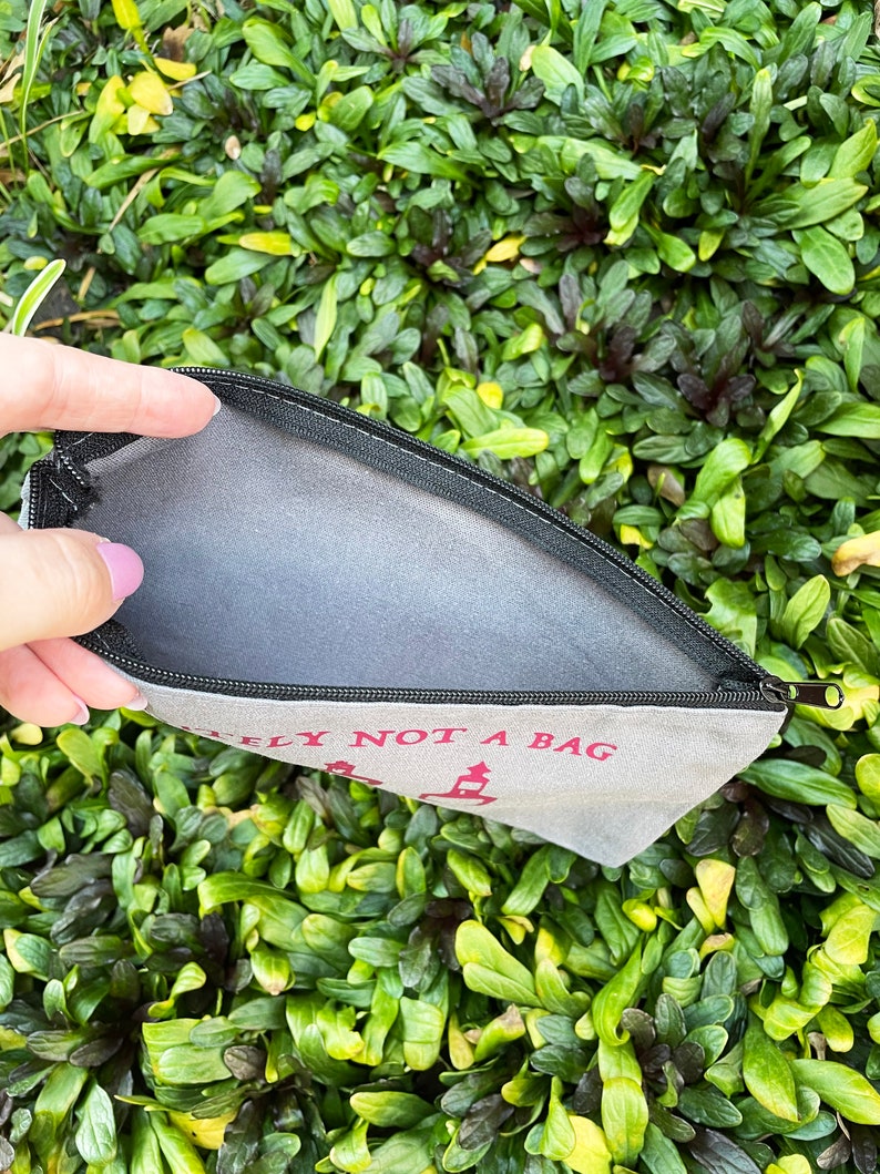 May include: A canvas zippered pouch with the text "DEFINITELY NOT A BAG" in red. The pouch is open, revealing a gray interior. The background is a dense bed of green foliage.
