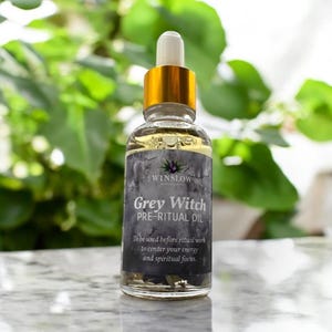 May include: A clear glass bottle with a gold cap and dropper, labeled "Grey Witch PRE-RITUAL OIL" by Winslow. The label is dark gray with white text. The bottle contains a light yellow liquid and is set against a blurred green background.
