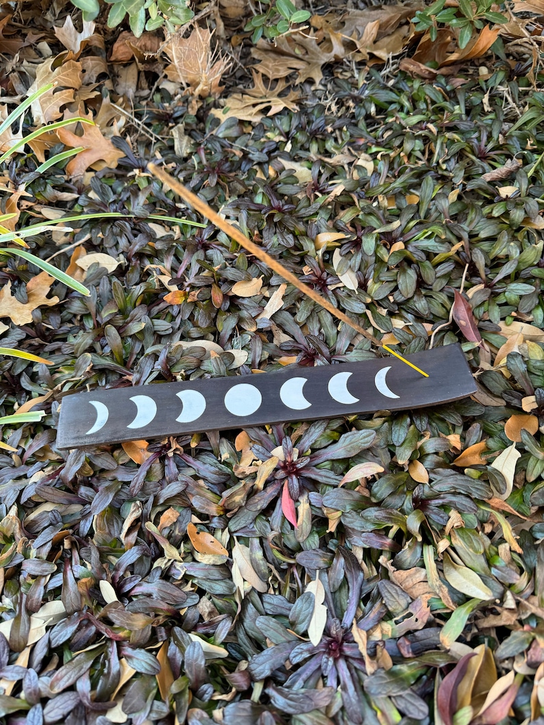 Puede incluir: Un porta incienso de madera negra con un dise&ntilde;o de fases lunares blancas. El porta incienso est&aacute; sentado en una cama de hojas marrones y verdes.