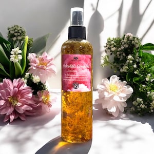 May include: A clear glass bottle with a gold liquid and flower petals inside. The bottle has a black spray nozzle and a white label with the text "Aphrodite's Command" and "Intention Spray" in black lettering. The bottle is sitting on a white surface with pink and white flowers.