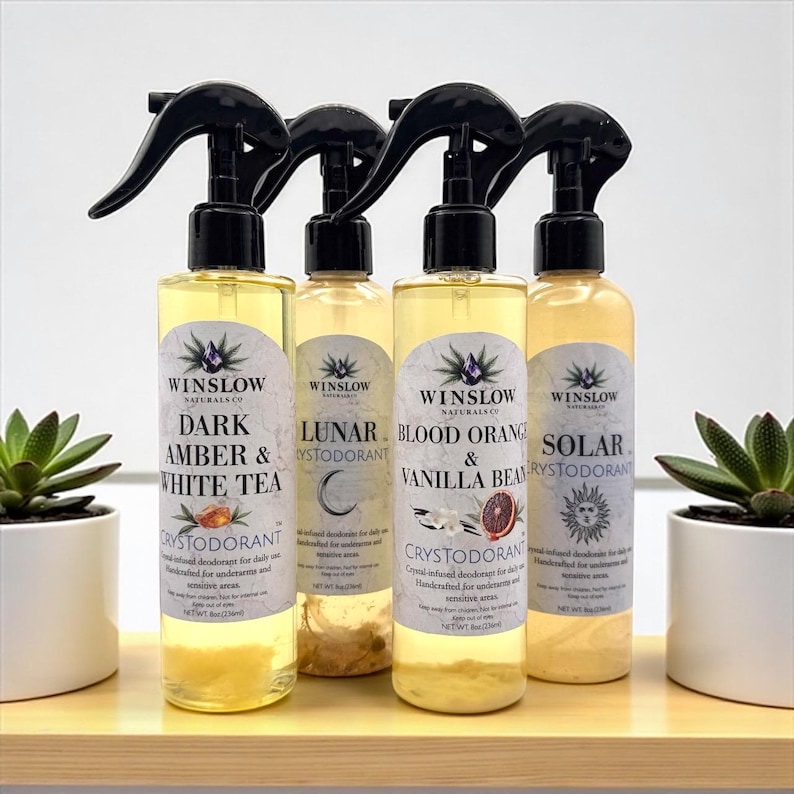 May include: Four clear spray bottles of crystal-infused deodorant, each with a black spray nozzle. The labels read "Dark Amber & White Tea", "Lunar", "Blood Orange & Vanilla Bean", and "Solar". The bottles are on a wooden shelf with small potted plants on either side.