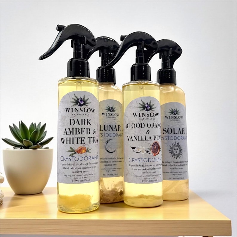 May include: Four bottles of Winslow Naturals Crystodorant, each with a black spray nozzle. The labels read "Dark Amber & White Tea", "Lunar", "Blood Orange & Vanilla Bean", and "Solar". The bottles are on a wooden shelf.