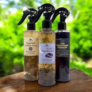 May include: Three spray bottles with black spray nozzles. The bottles contain clear liquid with herbs and labels that read "Hypnos Slumber Spray", "Evaki's Deep Sleep Spray", and "The Nightmare Barrier". The bottles are on a wooden surface.