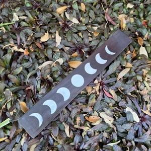 Puede incluir: Un porta incienso de madera marr&oacute;n oscuro con un dise&ntilde;o de lunas crecientes blancas. El porta incienso est&aacute; sobre un lecho de hojas verdes y marrones.