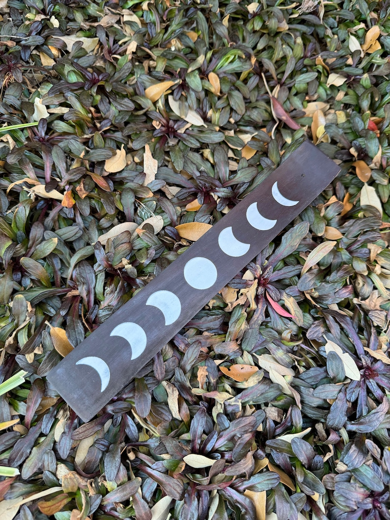 Puede incluir: Un porta incienso de madera marr&oacute;n oscuro con un dise&ntilde;o de lunas crecientes blancas. El porta incienso est&aacute; sobre un lecho de hojas verdes y marrones.