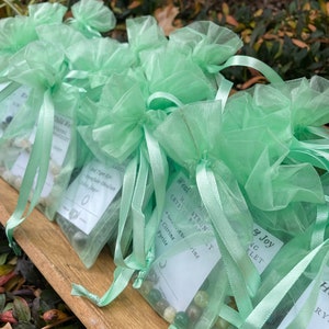 May include: Green organza bags filled with small stones and crystals. Each bag has a white tag with the text "Hand-Strung Crystal Bracelet" and a list of crystals, including Citrine, Pyrite, and Aventurine.
