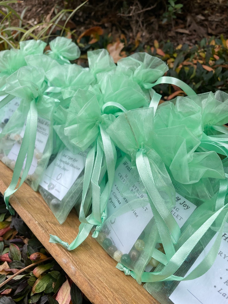 May include: Green organza bags filled with small stones and crystals. Each bag has a white tag with the text "Hand-Strung Crystal Bracelet" and a list of crystals, including Citrine, Pyrite, and Aventurine.