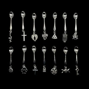 May include: Set of 15 silver teaspoons with various charms, including a cross, a snake, a crescent moon, a pentagram, a spider, an owl, a dragon, a heart, a key, a paw print, an ankh, a clover, a yin yang, a castle, and a swirl.