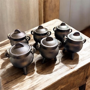 May include: A set of five small, dark, textured cast iron cauldrons, each featuring a rounded body, three short legs, two side handles with light-colored rings, and a matching lid with a central handle, arranged in a group on a wooden surface.